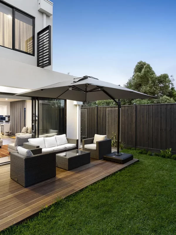 Modern two-story house with large sliding glass doors leading to an outdoor patio with a seating area and umbrella, surrounded by a well-manicured lawn and wooden privacy fence.