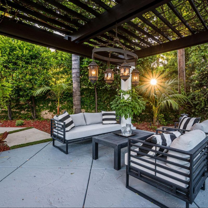 A patio featuring a black and white cushioned sofa set, surrounded by lush plants and sheltered by a black steel-framed acrylic cover.