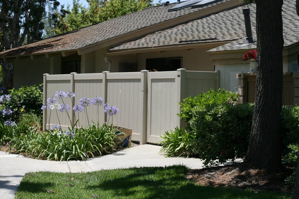 A beige fence in front of a house with purple flowers and greenery growing alongside it.