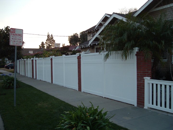 A residential street with a white fence and a 'No Parking' sign on the left side, and houses with palm trees on the right side.