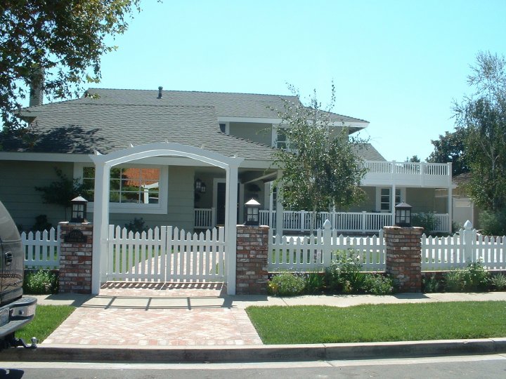 A one-story gray house with a white picket fence and brick pillars, archway entrance, and a small front yard with green grass and a tree.