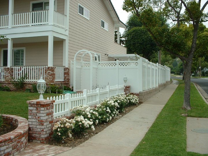 A beige two-story house with white trim and a white picket fence alongside a brick planter filled with white roses. A white vinyl privacy fence extends down the sidewalk, with a street and a tree in the background.