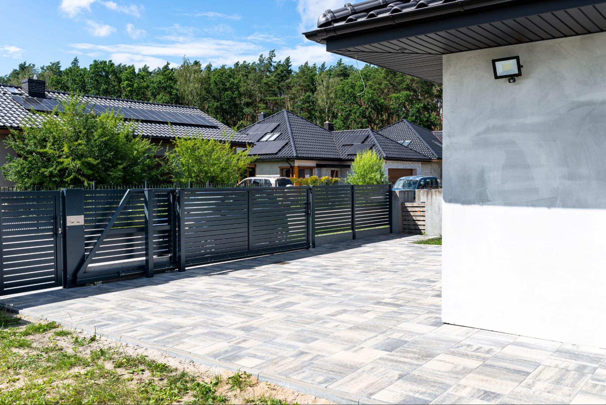 A modern anthracite panel fence with a coordinated sliding gate to the garage and a matching wicket gate, set against a backdrop of trees, houses, blue sky, and white clouds.