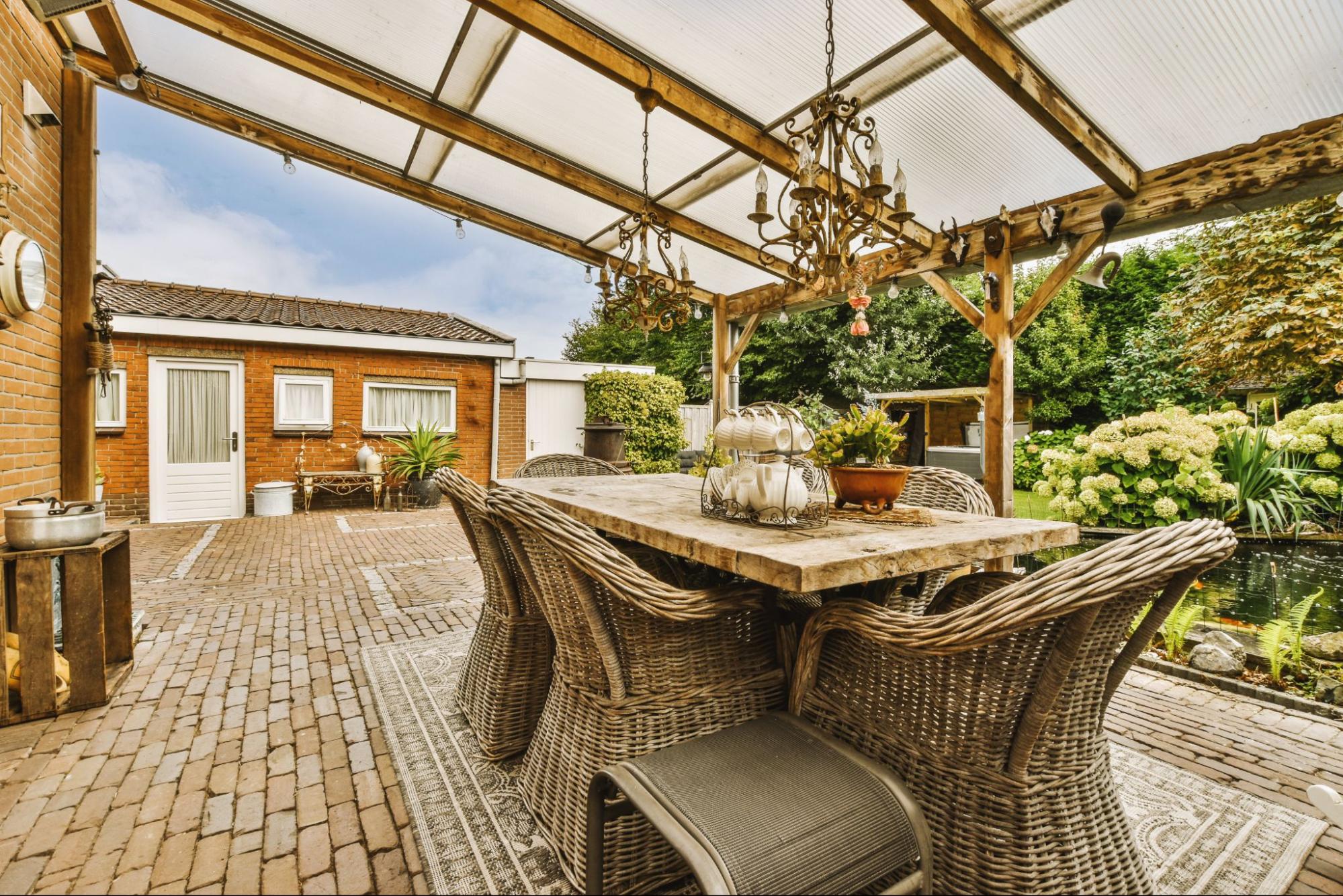 A neat patio covering an outdoor space with a rattan dining set facing a beautiful garden on the other side of a brick house; chandeliers under the ceiling add to the aesthetic appeal.