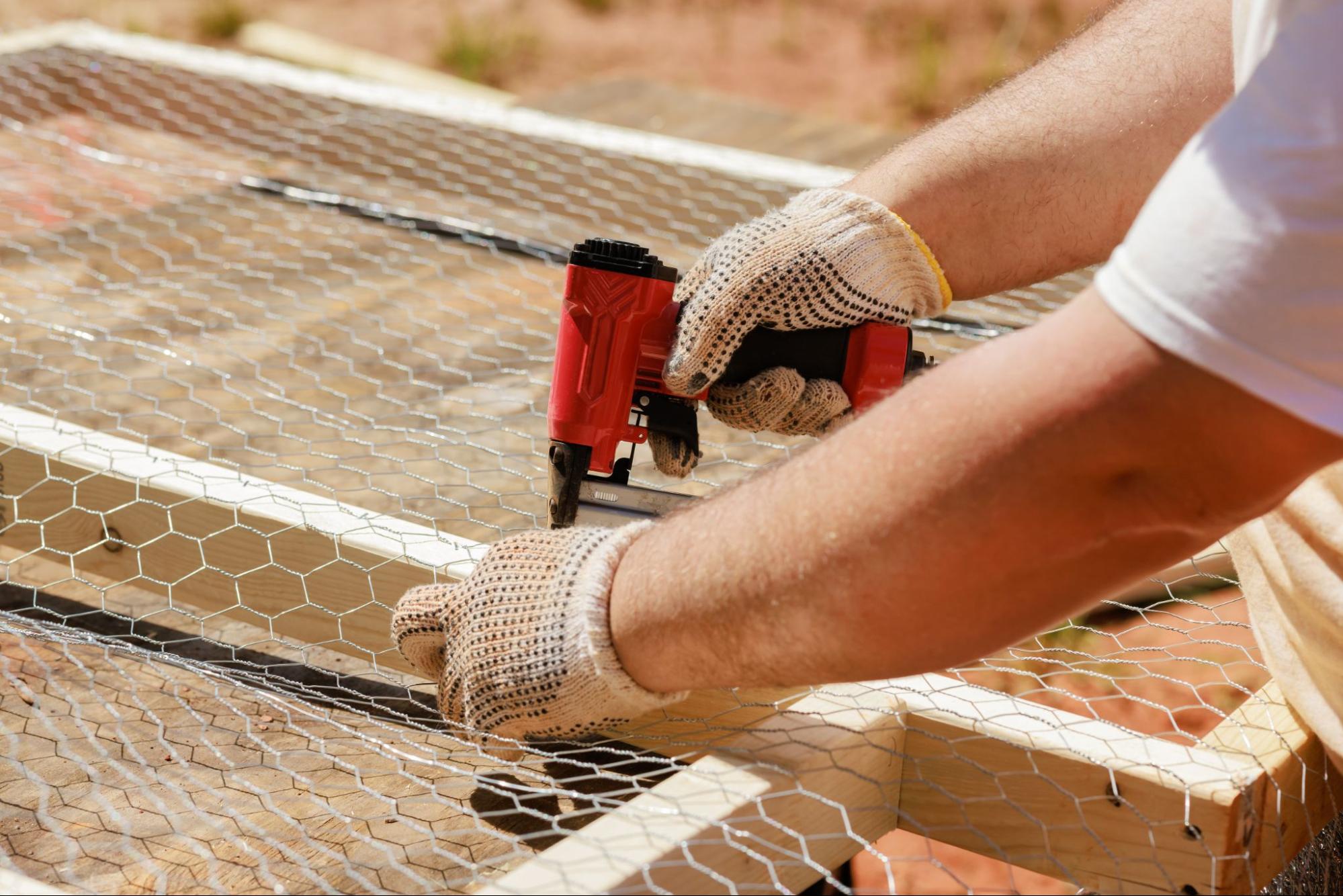 A man is nailing a metal mesh using a nail gun to a frame made of wood to make fences.