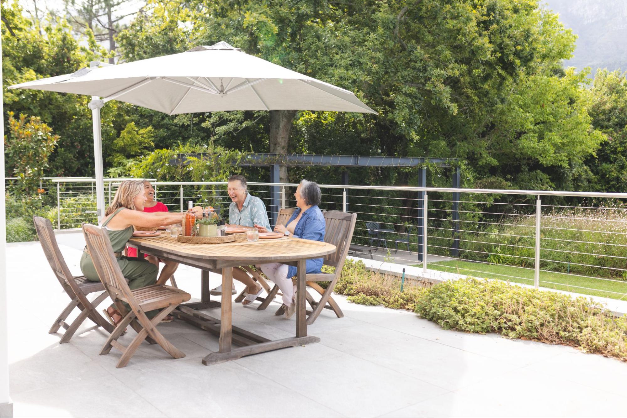 Three diverse senior female friends enjoying a meal outdoors, chatting happily and comfortably under a patio covering, with a spacious garden surrounded by lush trees in the background.