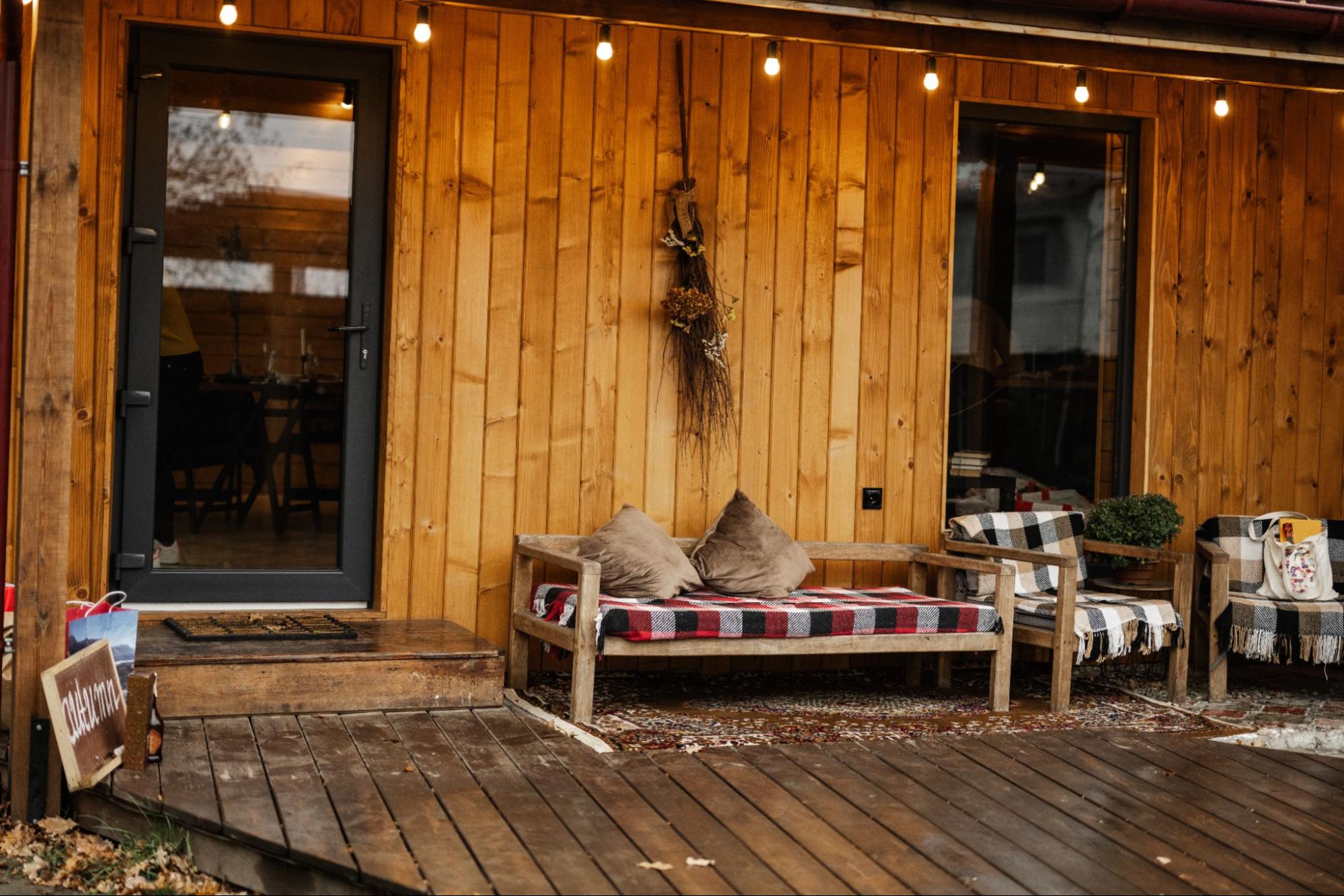A wooden couch with two brown throw pillows and wooden chairs with checkered-design cushions are set on the wooden deck of a cozy country house.