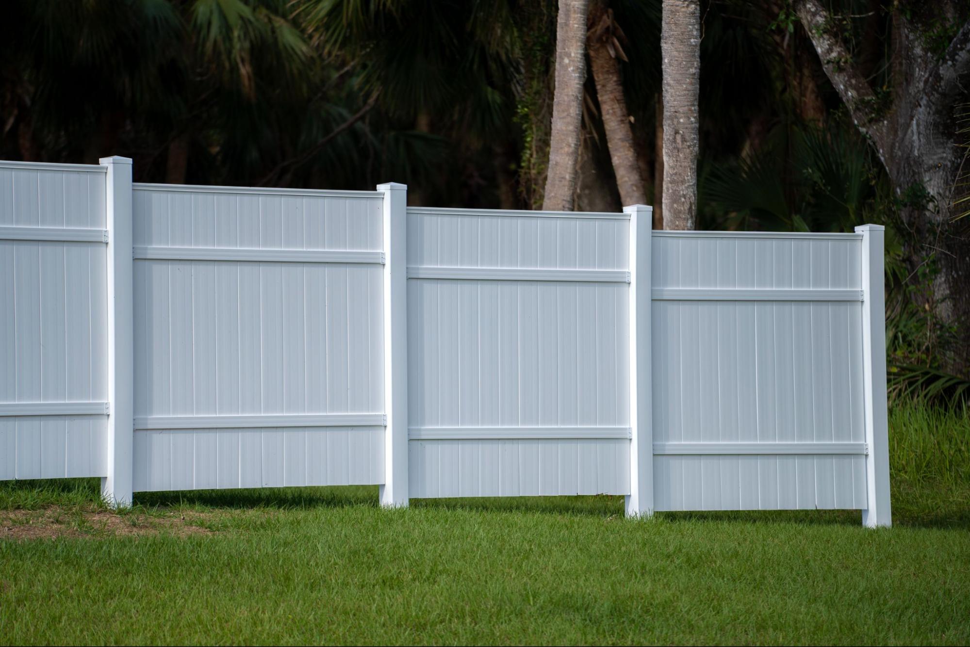 A white vinyl picket fence with trees in the background.