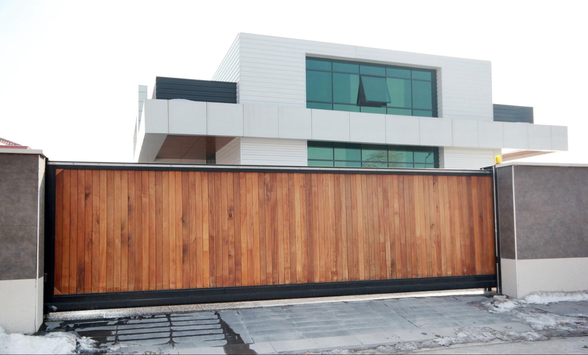 An automatic wooden entry door between concrete walls, with a white building featuring aluminum screen-covered glass windows in the background.