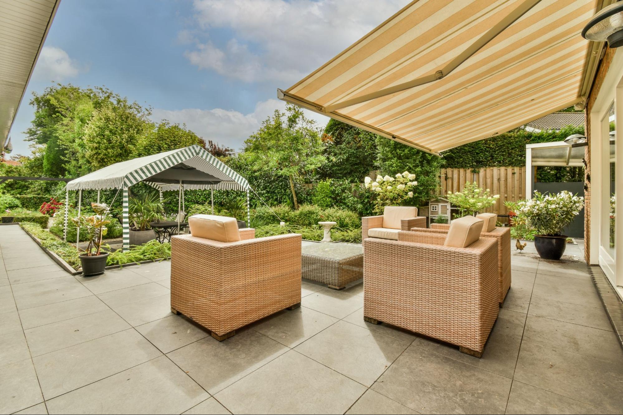 A beautiful patio with a cozy sitting area featuring a neat patio covering and facing a lush, vibrant garden filled with diverse plants and blooming flowers under the blue skies.