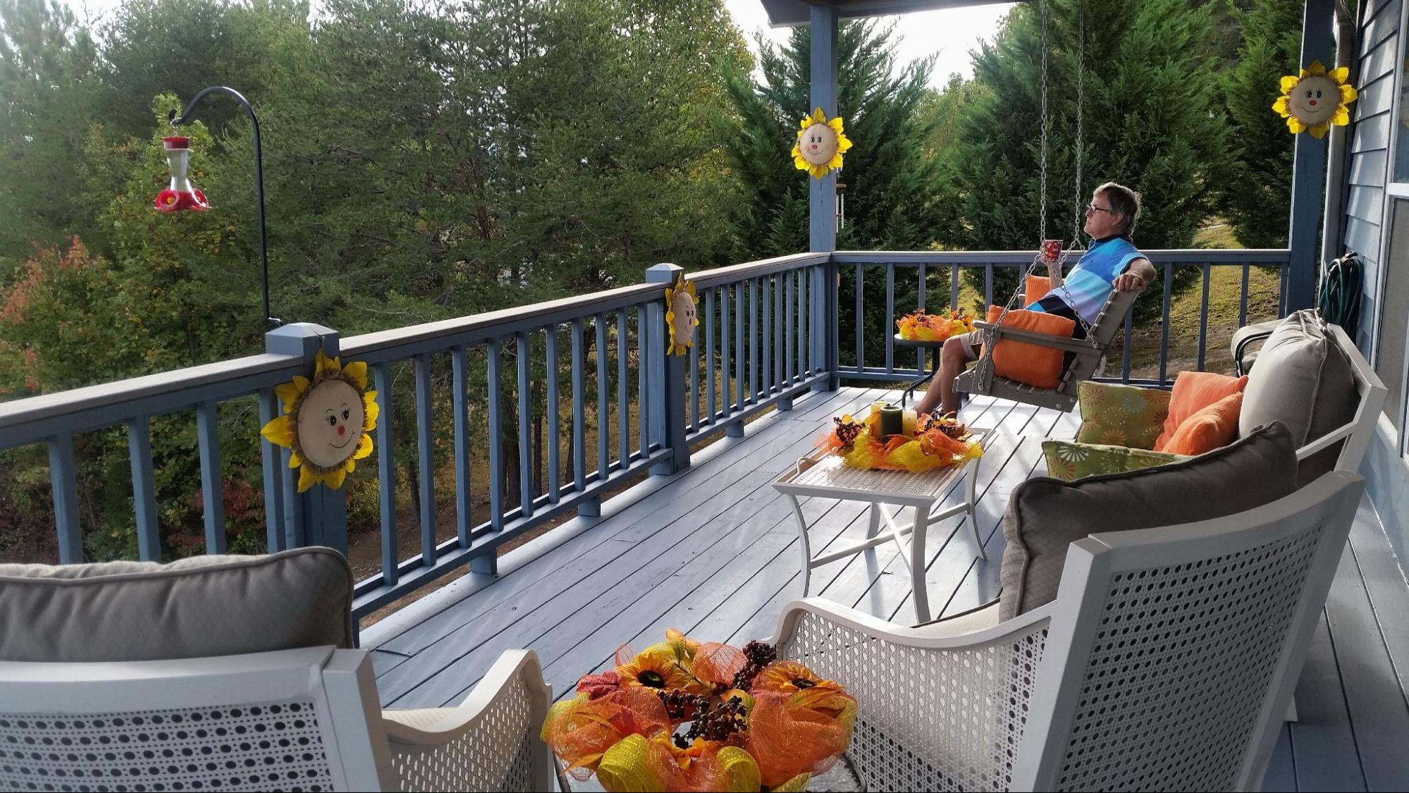 A man in a blue collared shirt sits on a swing with orange throw pillows, enjoying the morning mountain view and his first cup of coffee. The swing is at the far end of his elegant gray monochromatic deck adorned with yellow and orange decorative accents.