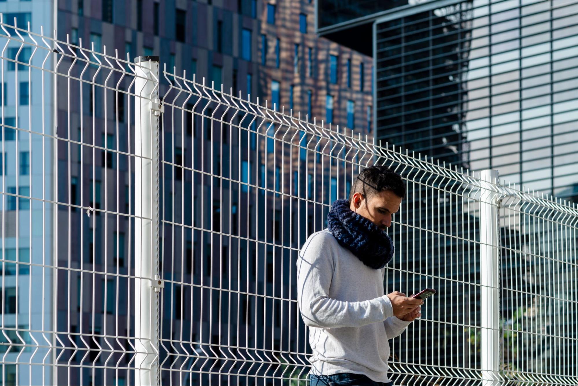 A man wearing a scarf leans on metallic fences.