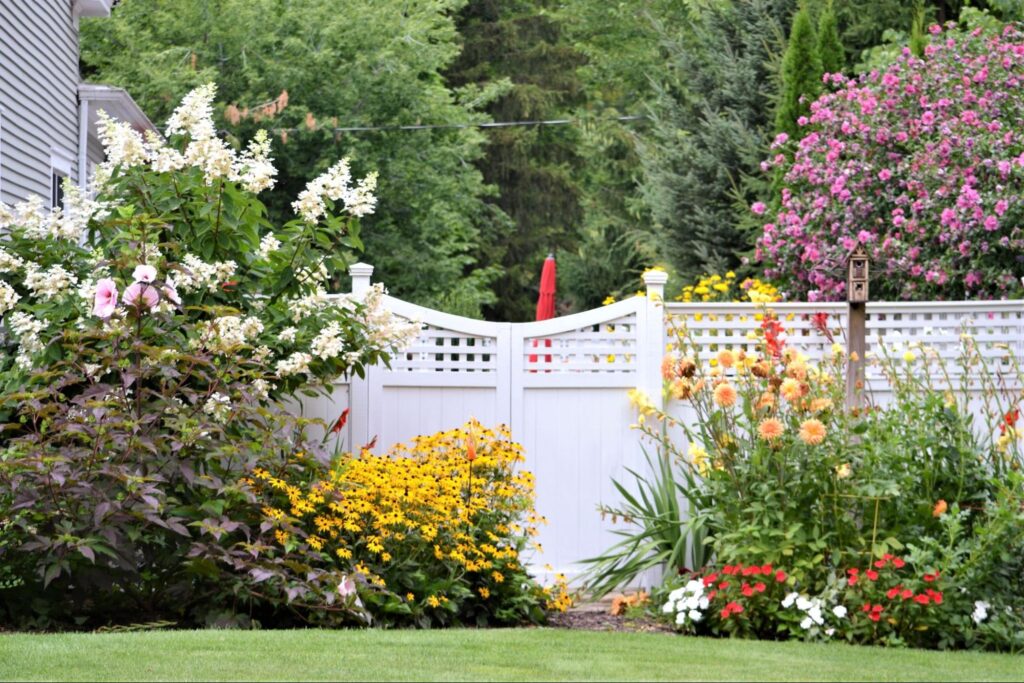 The white fence is surrounded by colorful and blooming flowers and trees.