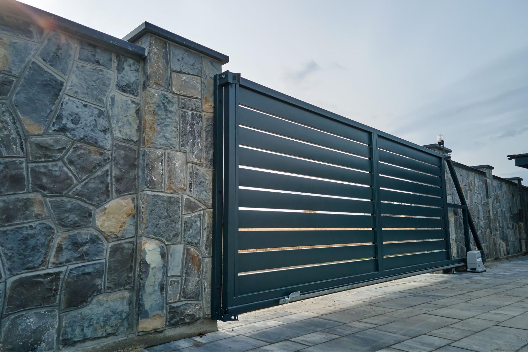 A wide automatic sliding gate with remote control installed in a high stone fence wall.