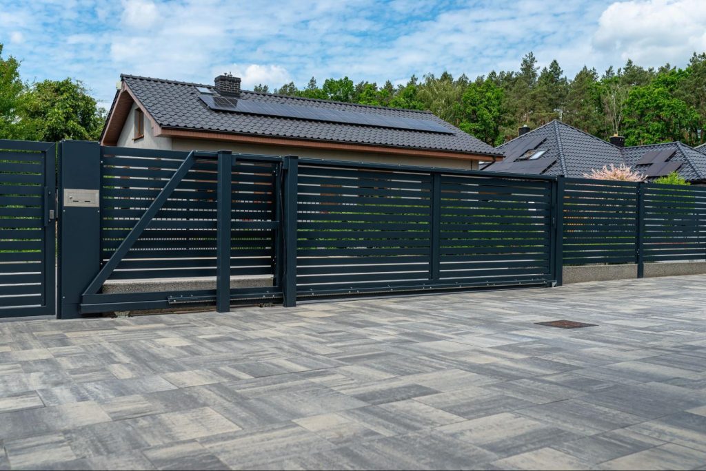 A modern anthracite-colored panel fence with a visible sliding gate leading to the garage and a slab driveway, with rows of houses visible behind it.