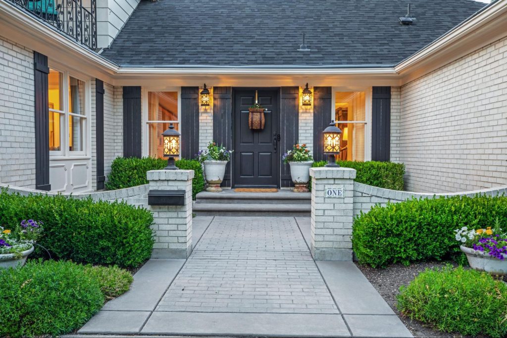 A white brick house with a gray roof, black door, and gray shutters features an exquisite front yard with manicured plants, two large white pots with flowering plants at the door, and a bowl-type pot with flowers, enhancing the property’s curb appeal.