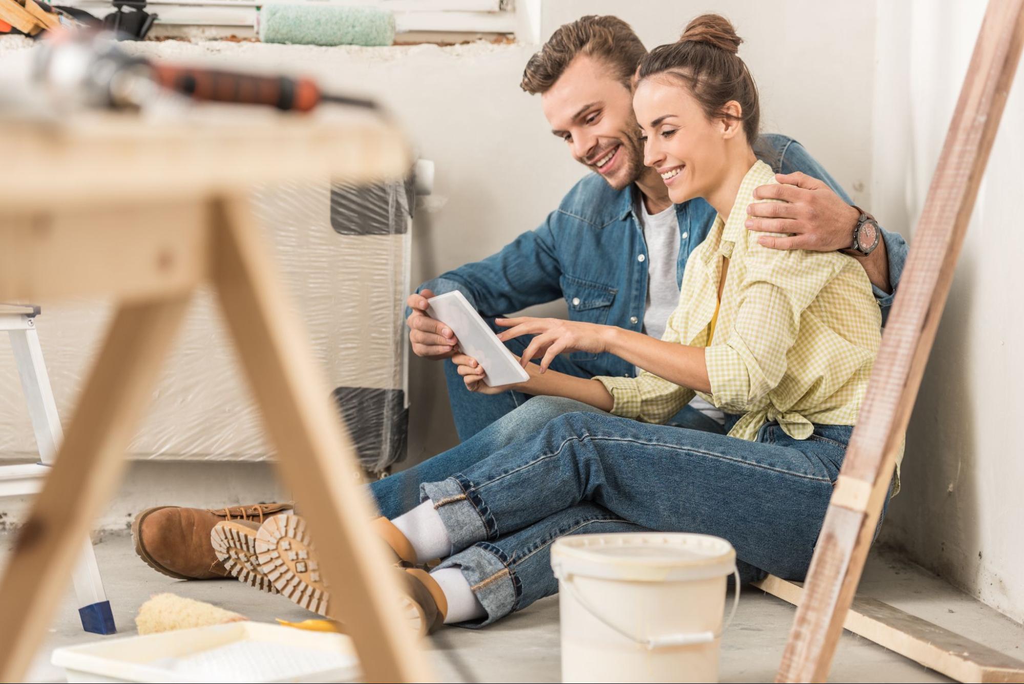 A happy young couple uses a digital tablet for guidance during repairs, with a plank of wood, a white bucket, and blurred tools on a desk in the foreground.