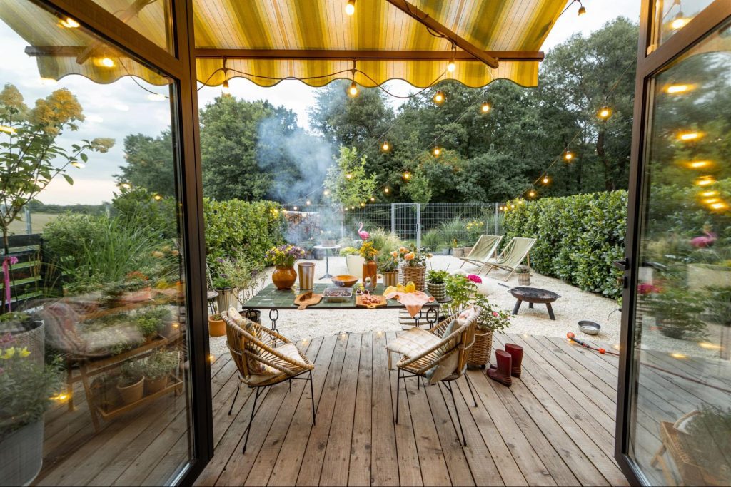 Wooden deck with a patio cover overhead with two open glass doors, a table, chairs, and twinkle lights.