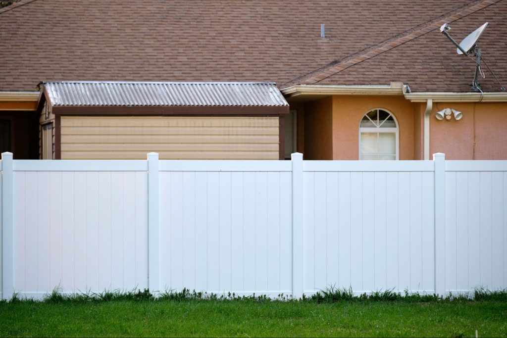 Vinyl fence next to grass, a shed, and a home. 