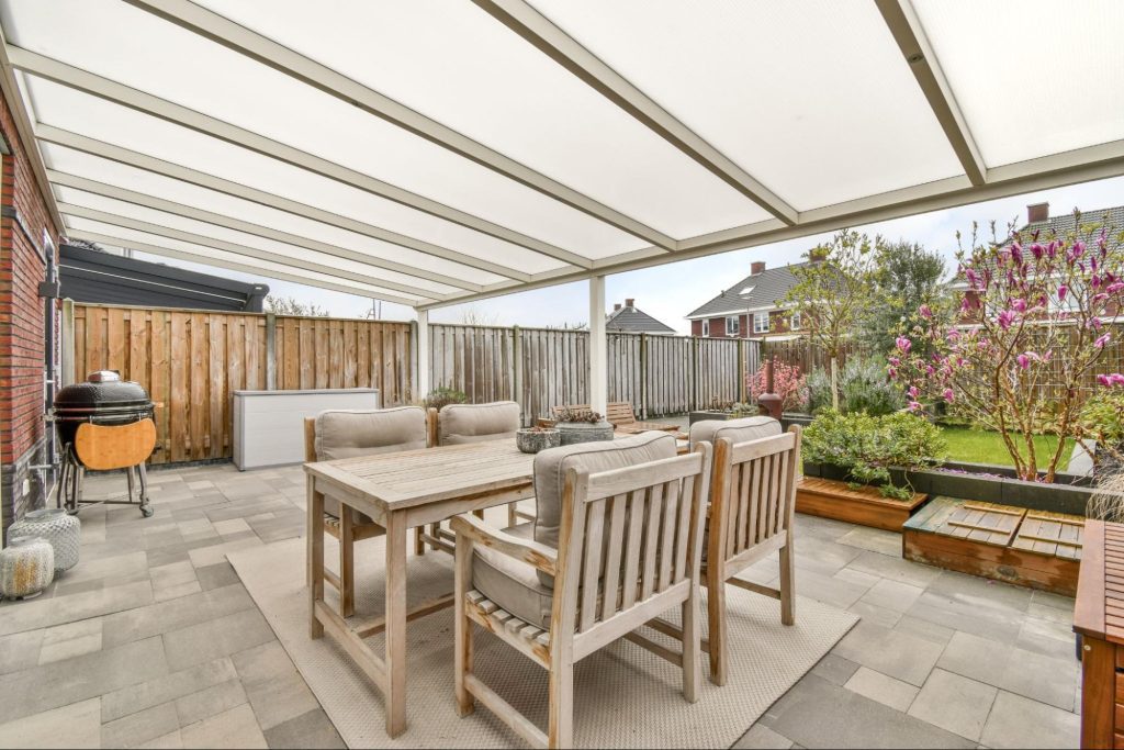 A well-designed covered patio with a cozy seating area facing a lush garden, featuring a portable BBQ grill on the side.