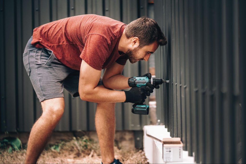 A man uses a cordless screwdriver to fasten screws on a black vinyl fence.