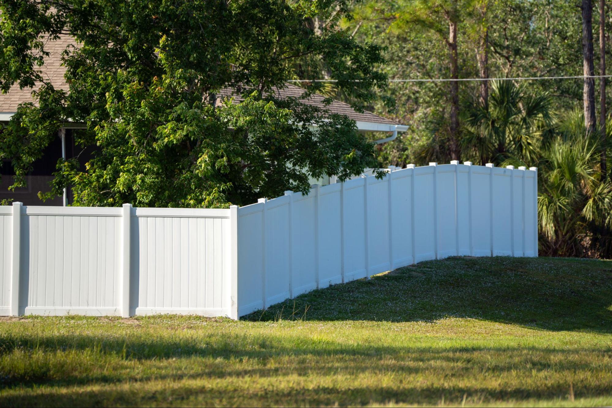 A white vinyl picket backyard fence encloses a vibrant green lawn, surrounded by tall, lush trees that create a tranquil and secure outdoor setting.