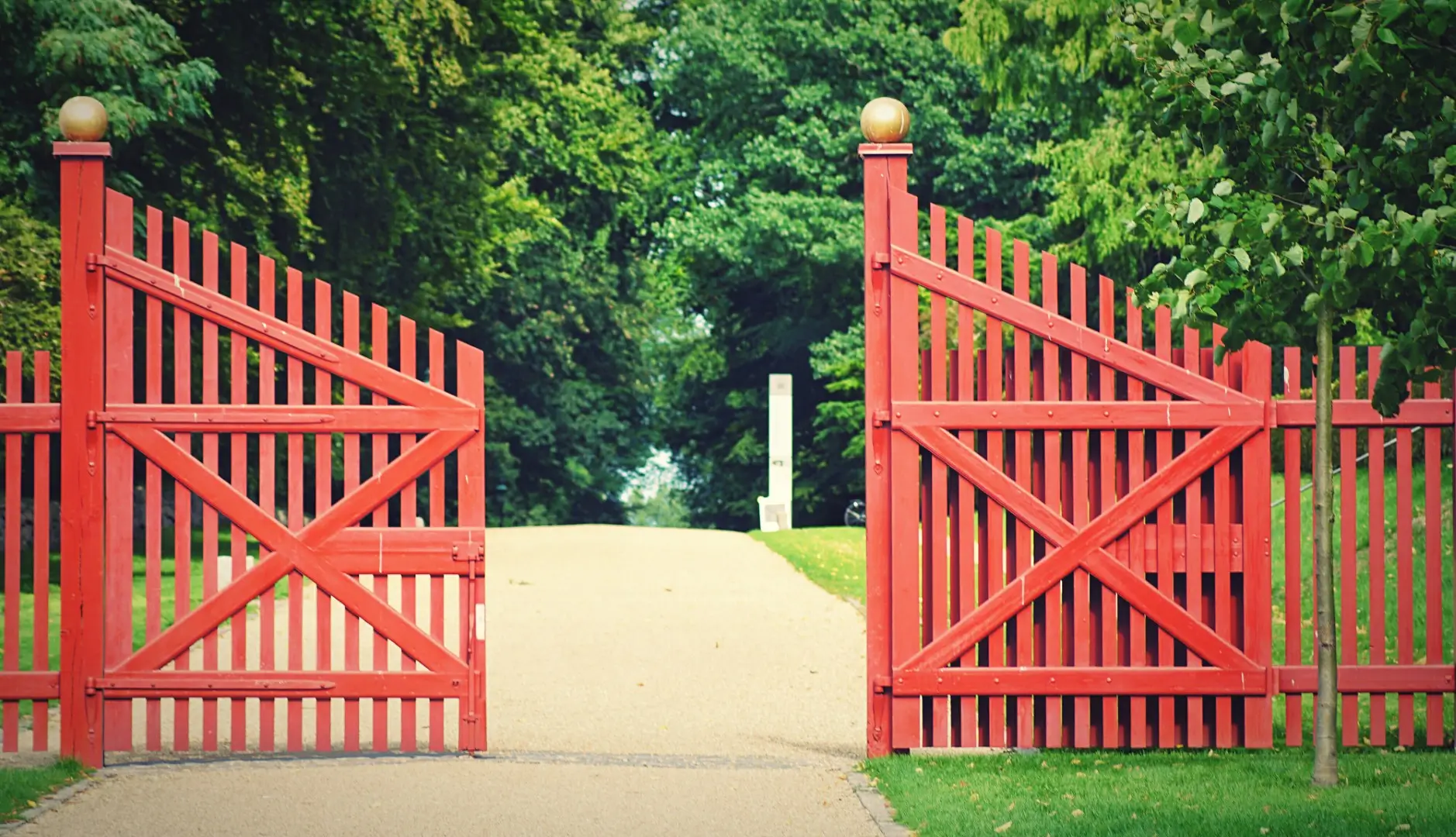 A half-open red fencing gate opening into a quiet park path.