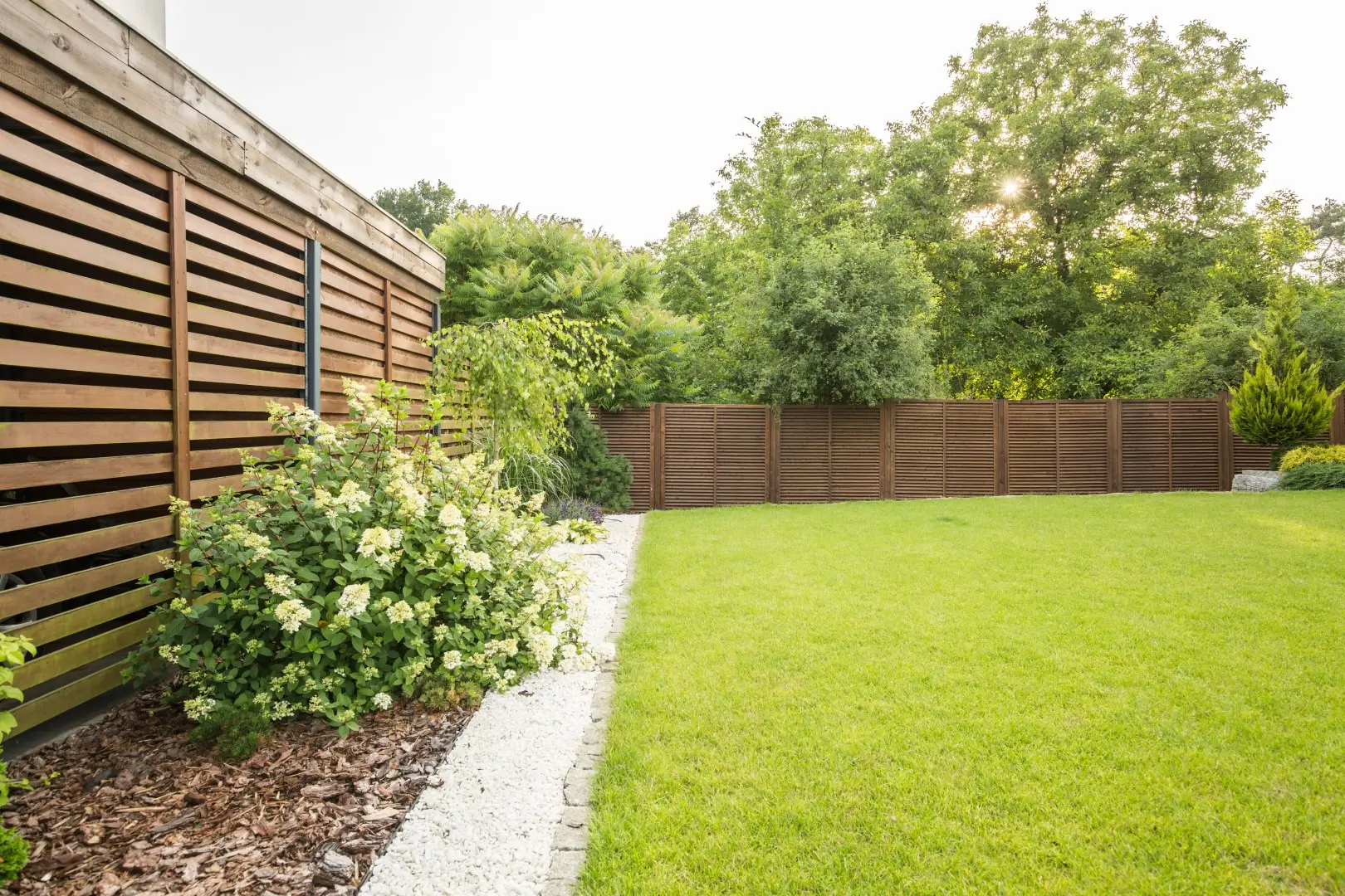 A neatly installed brown backyard fence with small garden trees, set in a serene yard with a house visible in the background.
