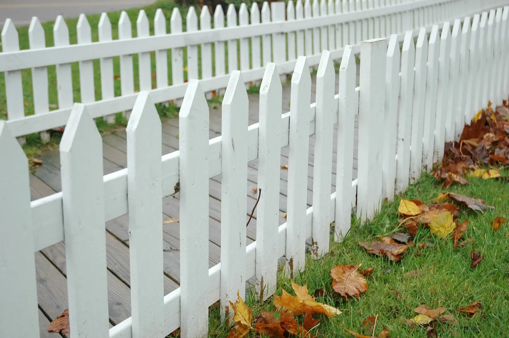 A white vinyl picket fence on a green lawn enclosing uneven property grounds.