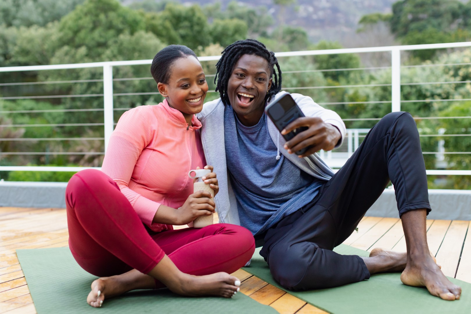 African American couple sitting on yoga mats on wooden deck holding water bottle and smartphone.