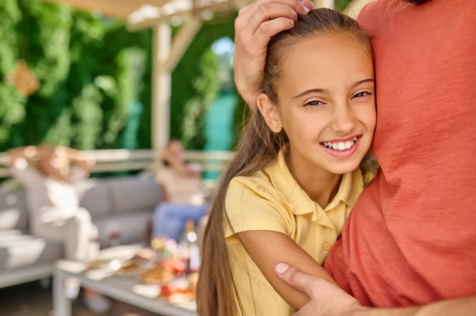 Man hugging his daughter under a vinyl patio cover with three people sitting on a couch in the background.