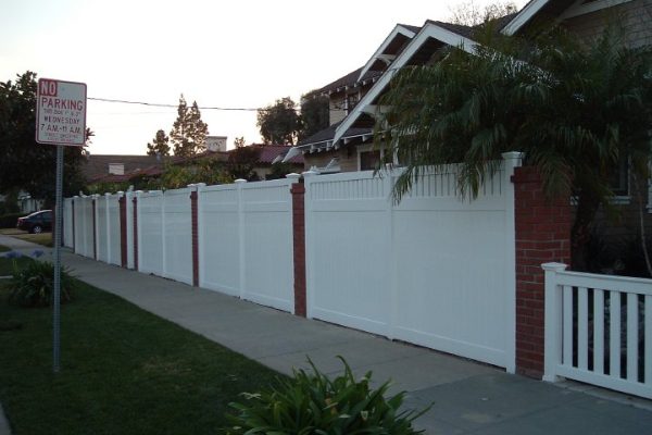 A residential street with a white fence and a 'No Parking' sign on the left side, and houses with palm trees on the right side.