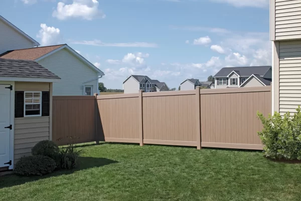 A beige fence in front of a house with purple flowers and greenery growing alongside it.