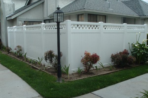 A white vinyl privacy fence with a decorative lattice top along the perimeter of a house's front yard, with a line of small shrubs planted in front and a black lamp post on the left side.