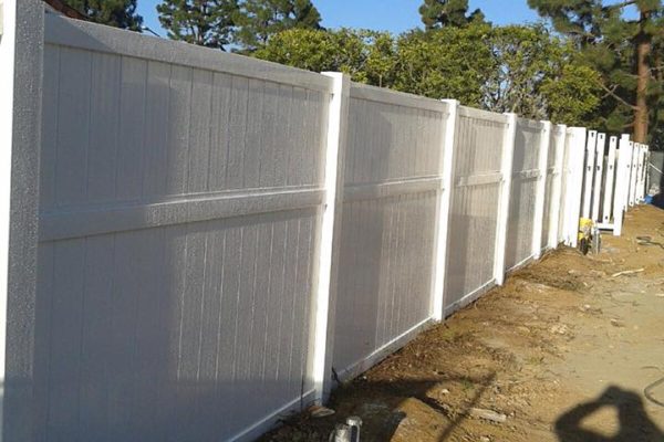 A white vinyl fence installed along a dirt path with trees and a blue sky in the background.