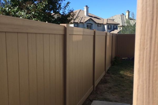 A beige wooden fence runs along the side of a yard, with a concrete path leading to a green lawn and the tops of houses visible in the background.