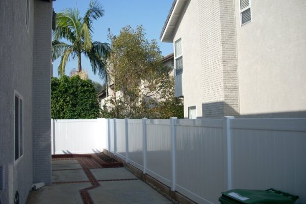 A narrow pathway between two beige houses with a white fence on one side and palm trees in the background.