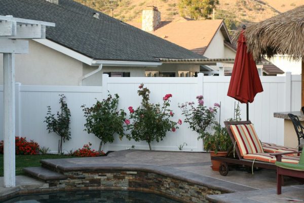 A well-manicured backyard with a white fence, blooming rose bushes, a pool with a stone edge, and a lounging chair with red and green stripes next to a closed red umbrella.