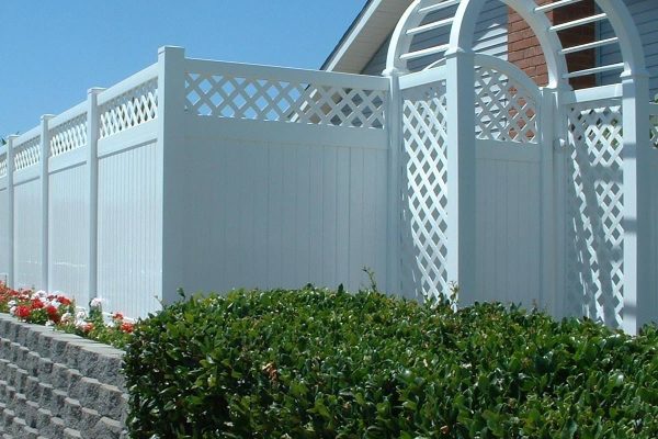 A white vinyl fence with a lattice pattern, an arched gate, and red and white flowers behind it, in front of a house with a brick chimney.