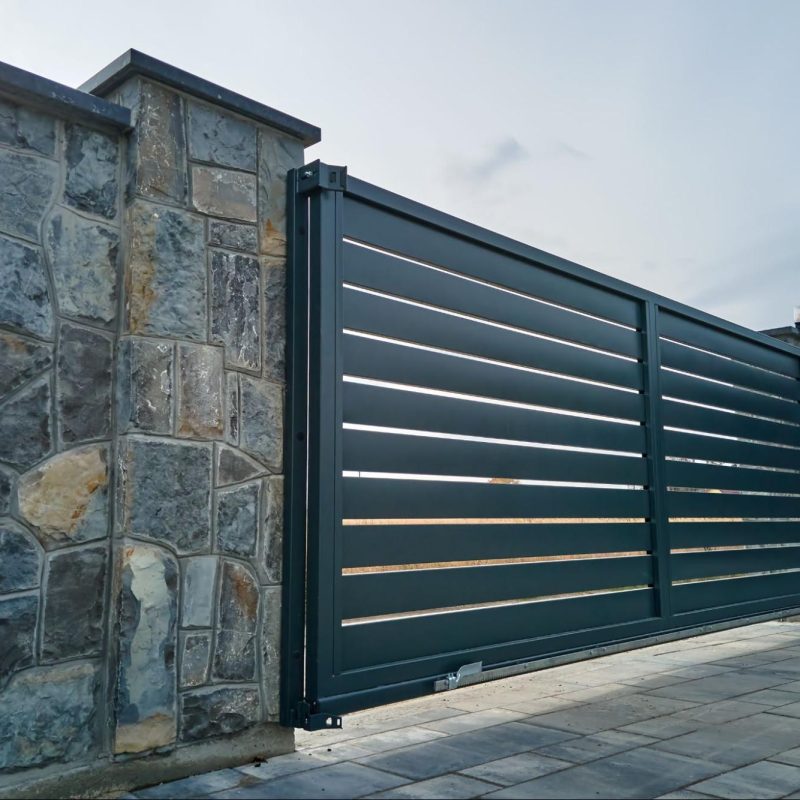 A wide automatic sliding gate with remote control installed in a high stone fence wall.