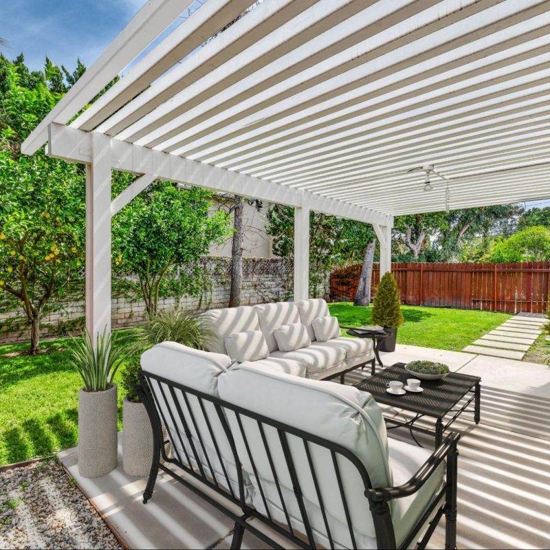 A sunny patio with a 3D-decorated house featuring a stylish vinyl patio cover with a slatted design for shade and elegance.