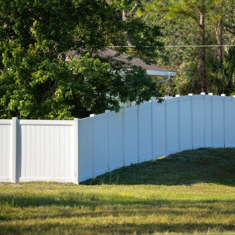 A white vinyl picket backyard fence encloses a vibrant green lawn, surrounded by tall, lush trees that create a tranquil and secure outdoor setting.