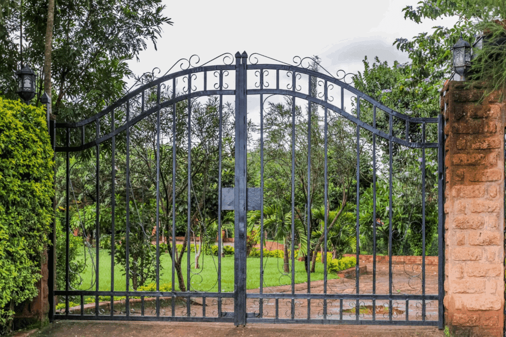 A modern black steel gate with clean lines, set outdoors against a green, well-decorated home exterior