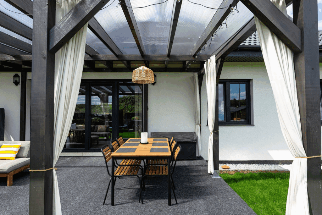 Patio with exposed wooden beams and a polycarbonate roof attached to the rear of a modern house.